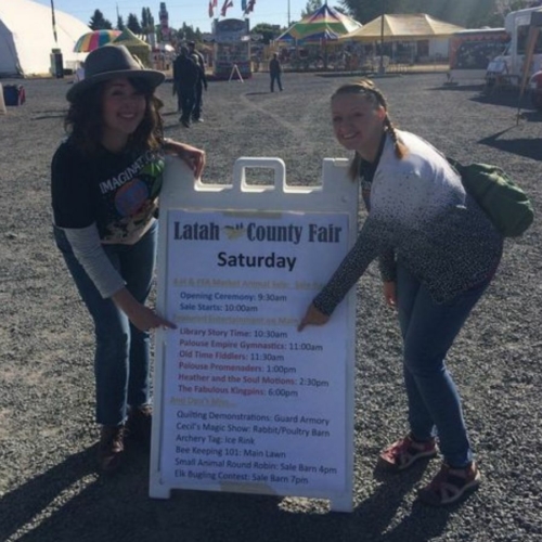 Miss Bailey and Miss Stacie are standing to either side of the Latah County Fair Saturday schedule, pointing at their slot for Library Storytime at 10:30 am on the main stage.
